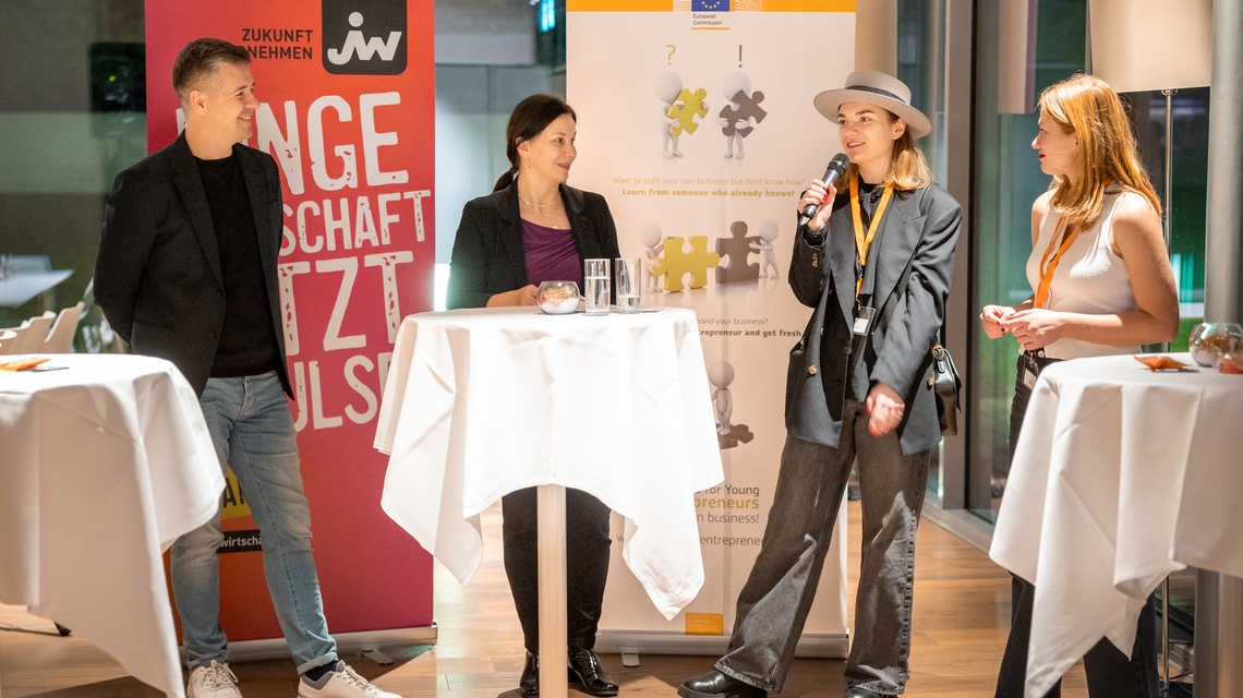 Zu sehen sind Personen, eine davon hat ein Mikrofon in der Hand. Im Hintergrund sind Roll-ups, auf einem steht 