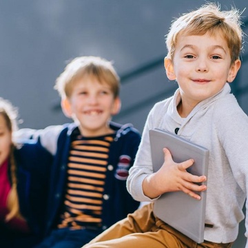 Fröhliches Kind mit Buch sitzt auf einer Bank und blickt in die Kamera während im Hintergrund zwei weitere glückliche Kinder sitzen