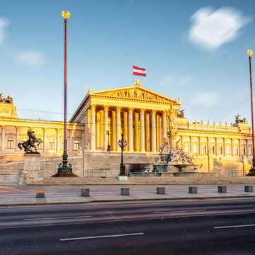 Frontale Aufnahme des Parlaments in Wien mit einer österreichischen Flagge am Dach sowie einem Brunnen vor dem Gebäude. Der Himmel ist leicht bewölkt. Die Sonne scheint auf Teile des Gebäudes. Nirgends sind Menschen