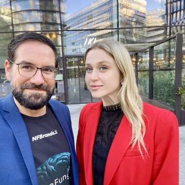 Porträt von zwei nebeneinander stehenden Personen vor einem Gebäude auf einem asphaltierten Platz. Die linke Person hat kurze, dunkle Haare und trägt Brille. Die rechte Person hat offene, helle Haare und trägt ein dunkles Oberteil