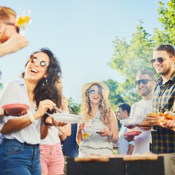 Mehrere Personen in sommerlicher Kleidung und mit Sonnenbrillen stehen in einem Garten. Teilweise halten sie Teller. Eine Person trägt eine Schürze und hält eine Grillzange. Die Personen blicken sich an. In der Mitte steht ein Grill