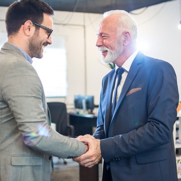 Zwei einander anlächelnde Personen in Anzügen schütteln sich die Hände, im Hintergrund verschwommen offener Büroraum
