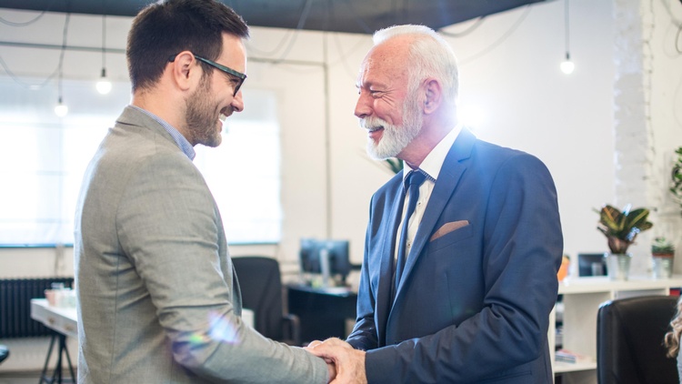 Zwei einander anlächelnde Personen in Anzügen schütteln sich die Hände, im Hintergrund verschwommen offener Büroraum
