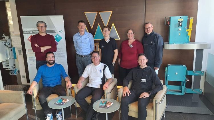 Gruppe von sieben Personen in einem Firmenfoyer, einige sitzen auf Sesseln, andere stehen dahinter. Im Hintergrund sind ein Firmenlogo 