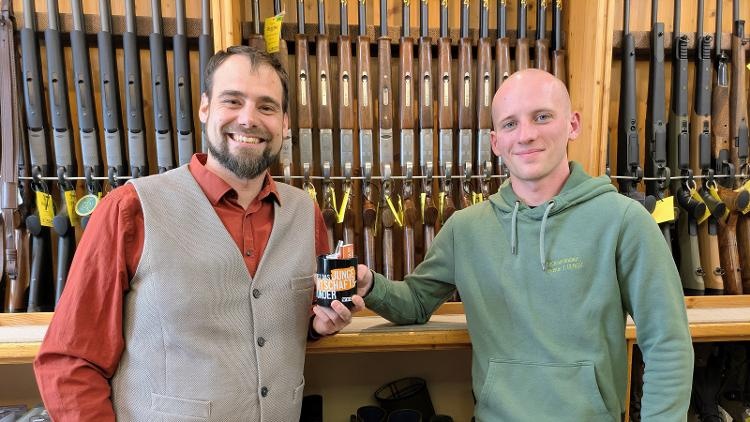 Zwei Personen stehen in einem Geschäft vor einer Wand mit Jagd- und Sportgewehren und halten gemeinsam eine schwarzes Häferl mit Aufdruck 