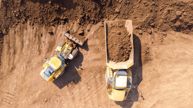 Aufnahme eines Baggers sowie eines Fahrzeuges mit einem großen Container aus der Vogelperspektive. Im Container ist Erde genauso wie auf der Schaufel des Baggers. Neben Bagger und Fahrzeug ist ein großer Haufen Erde