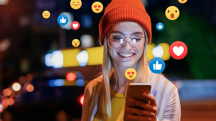 Lächelnde Person mit Brille und Haube hält ein Smartphone in der Hand während verschiedene Social Media Icons wie Herzen, Daumen und Gesichter aufsteigen