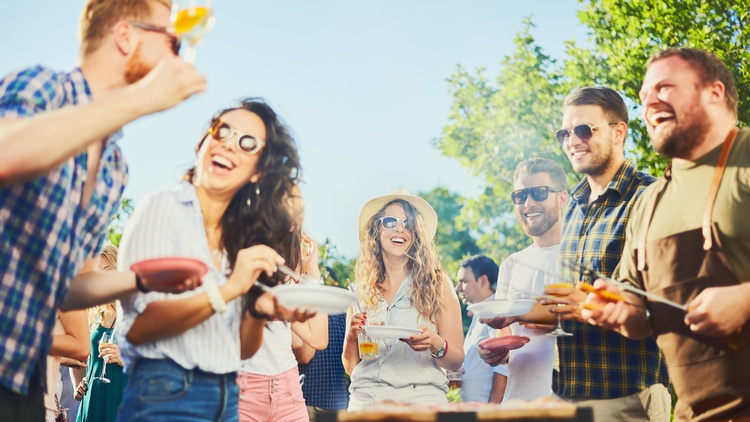 Mehrere Personen in sommerlicher Kleidung und mit Sonnenbrillen stehen in einem Garten. Teilweise halten sie Teller. Eine Person trägt eine Schürze und hält eine Grillzange. Die Personen blicken sich an. In der Mitte steht ein Grill