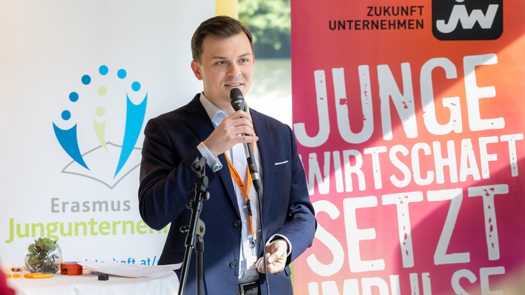 Eine Person mit Mikrofon in der rechten Hand steht auf einer Bühne. Im Hintergrund ist ein Roll-Up mit dem Schriftzug Junge Wirtschaft setzt Impulse und ein Plakat mit dem Schriftzug Erasmus Jungunternehmer
