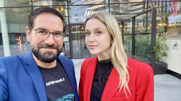 Porträt von zwei nebeneinander stehenden Personen vor einem Gebäude auf einem asphaltierten Platz. Die linke Person hat kurze, dunkle Haare und trägt Brille. Die rechte Person hat offene, helle Haare und trägt ein dunkles Oberteil
