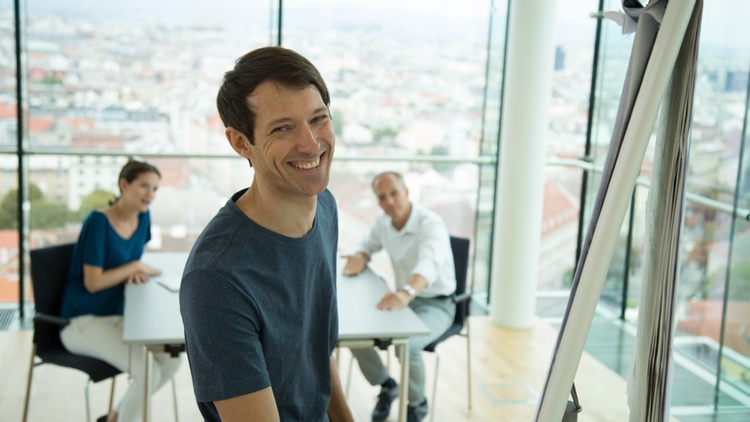 Ein Mann präsentiert etwas an einem Flipchart, im Hintergrund sitzen zwei Zuschauer.