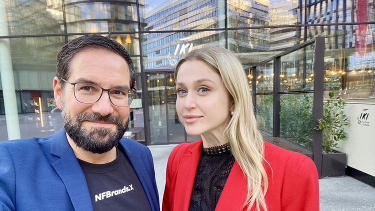 Porträt von zwei nebeneinander stehenden Personen vor einem Gebäude auf einem asphaltierten Platz. Die linke Person hat kurze, dunkle Haare und trägt Brille. Die rechte Person hat offene, helle Haare und trägt ein dunkles Oberteil
